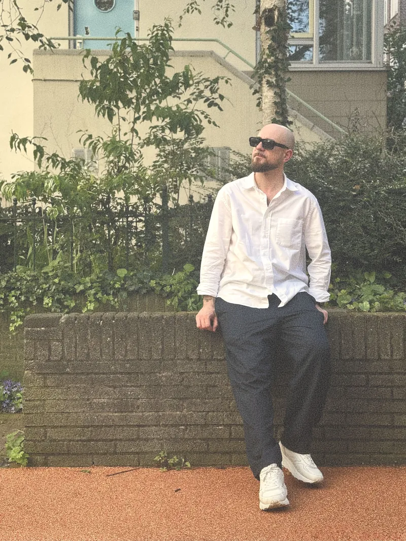 Alessandro sitting outdoors beside trees and plants, wearing sunglasses and a white shirt.